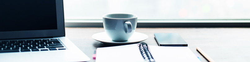 A desk with a laptop, open notebook and coffee cup sitting on top of it.